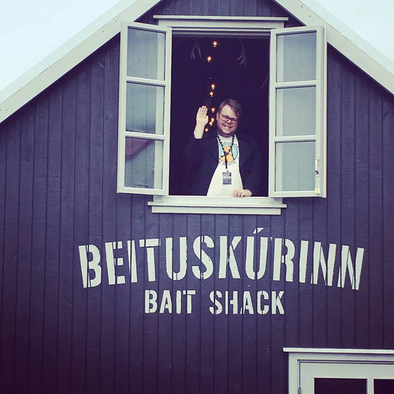 Manneskja lyftir glasi í loftglugganum fyrir ofan skilti Beituskúrsins